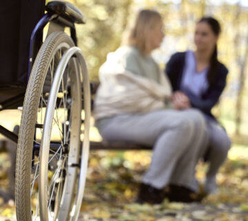 Rollstuhl mit zwei Menschen auf Bank im Hintergrund © Westend61 / gpointstudio